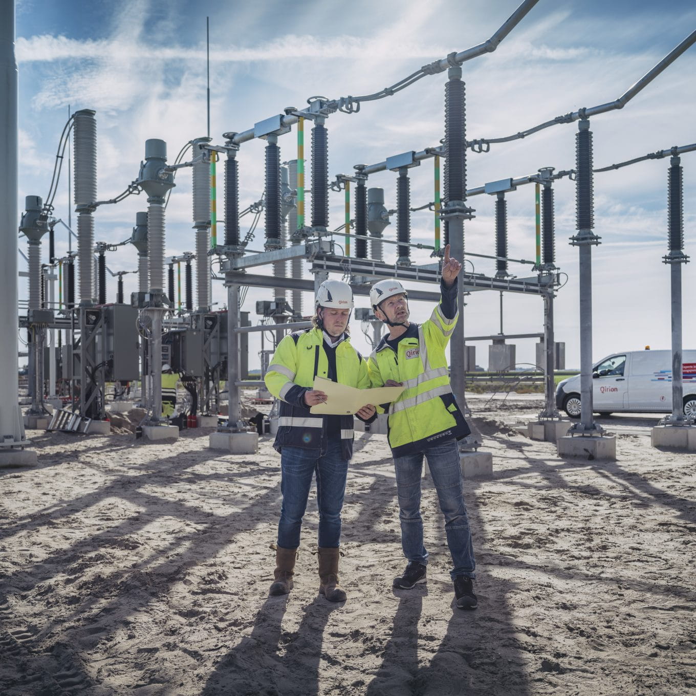 Twee monteurs in veiligheidskleding bekijken een bouwtekening bij een hoogspanningsstation en wijzen naar de installatie.